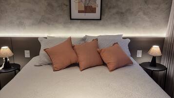 Cama bem arrumada com um edredom branco, duas almofadas castanhas e um cartão sobre a cama. Há uma obra de arte emoldurada na parede acima da cama.