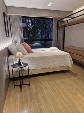 A modern bedroom with a large bed, a bedside table with a lamp, and a view of trees through the window.