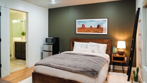 Un dormitorio con cama, una mesita de noche con lámpara, un cuadro en la pared y un cactus en el suelo.