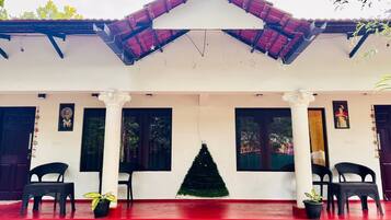 Un edificio con tejado rojo, paredes blancas y un árbol de Navidad verde en el centro. Hay dos sillas negras y dos plantas en macetas sobre el suelo rojo.