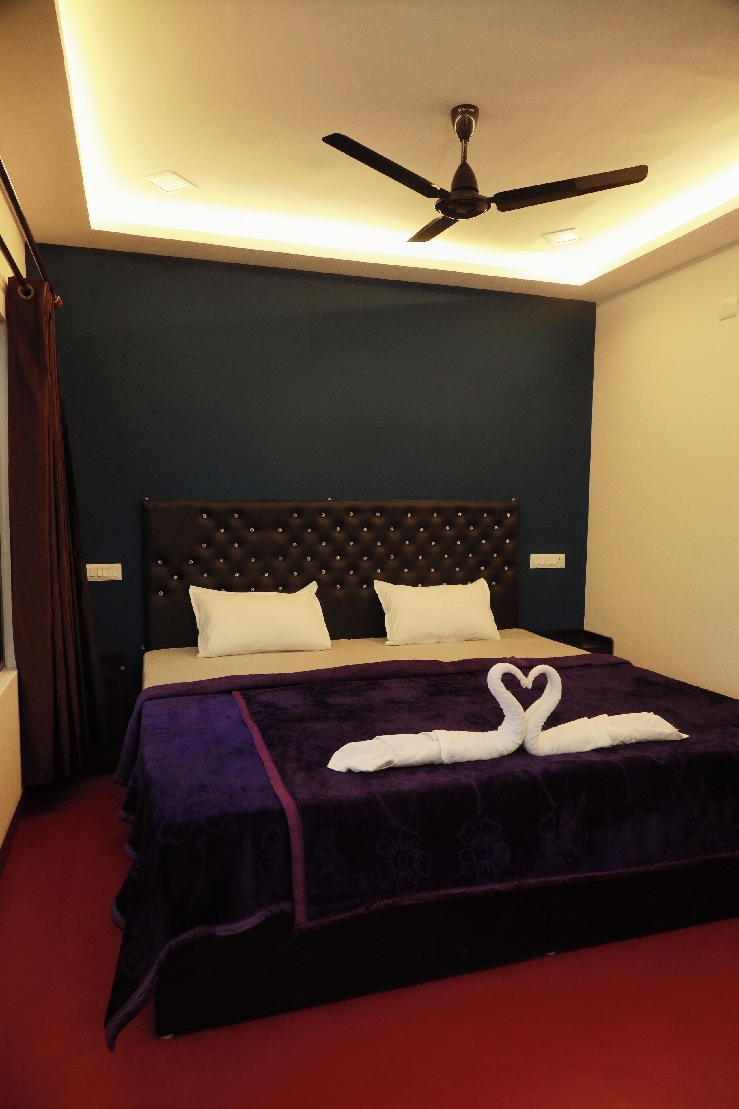A bedroom with a tufted headboard, a bed with white pillows, a purple bedspread, and a heart-shaped towel arrangement on the bed.