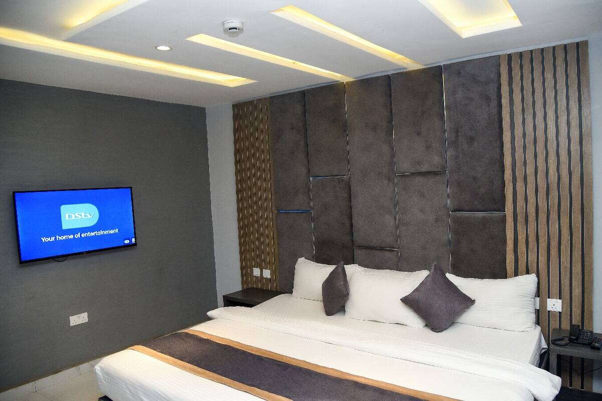Une chambre d’hôtel moderne dotée d’un grand lit, d’un téléviseur fixé au mur diffusant la chaîne « DSTV, votre source de divertissement », et d’un panneau mural au design original.