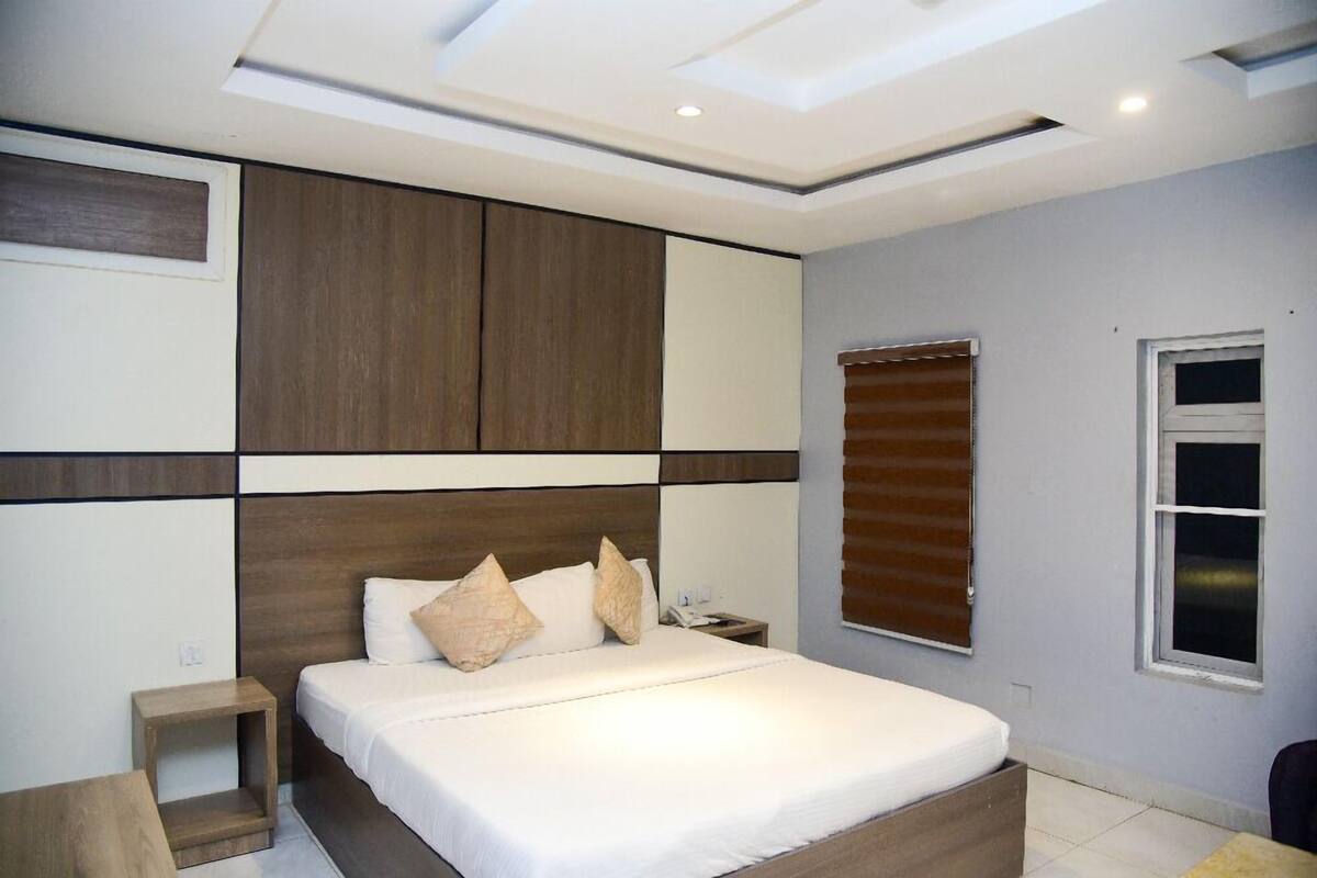 Une chambre à coucher avec un grand lit, une table de chevet, une tête de lit en bois, une fenêtre avec des stores et un plafond doté d’un éclairage encastré.