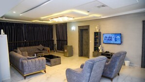 A modern living room with a sectional sofa, a coffee table, and two armchairs. A flat-screen TV is mounted on the wall, and there is a small table with a vase and a statue. - Hotel De Alommah Kubwa Abuja Nigeria (Abuja)