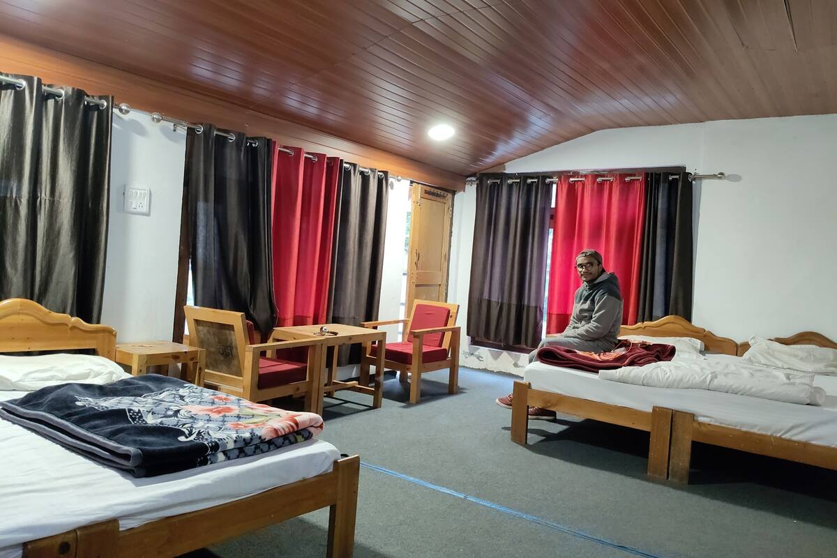 Une chambre d’hôtel avec deux lits, une table en bois, une chaise et une personne assise sur l’un des lits.