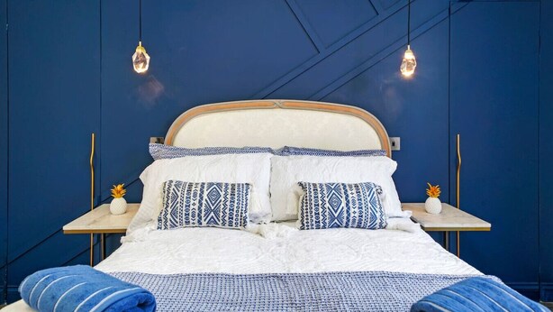 Ein Bett mit gemusterten Kissen, einer weißen Decke und blauer Bettwäsche, vor einer blauen Wand mit geometrischem Muster.