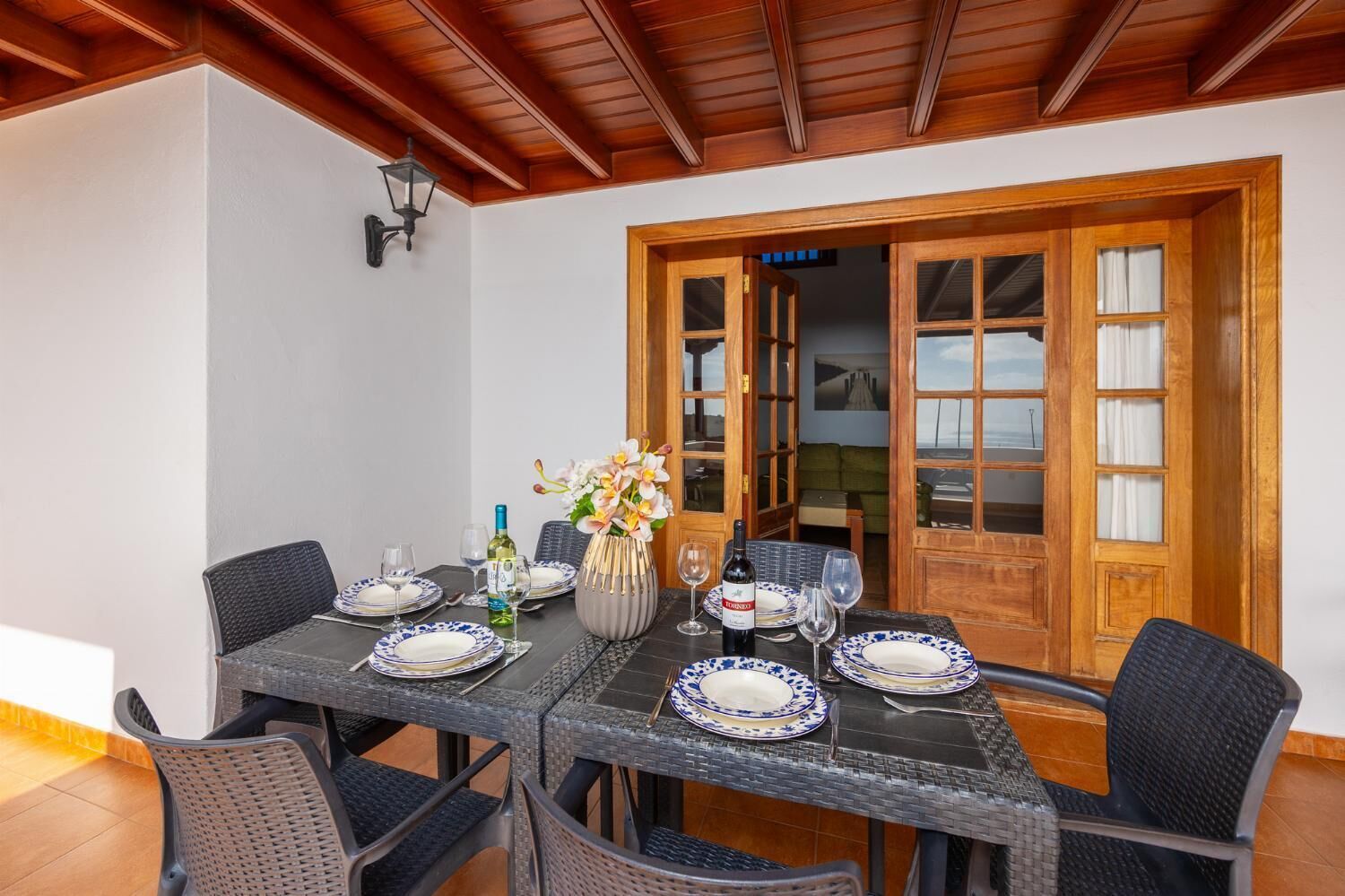 A dining area with a table set for six, surrounded by wicker chairs, a vase with flowers, and a bottle of wine.