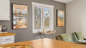 A dining area with a wooden table, chairs, and a window with a view of trees. There are two framed posters on the wall. - Stylish & Spacious Home | Hot Tub & 3-Season Deck (Douglas)