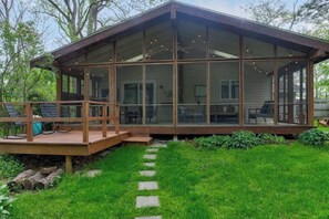 A wooden deck with outdoor furniture, a glass-enclosed structure, and a stone pathway. - Stylish & Spacious Home | Hot Tub & 3-Season Deck (Douglas)