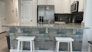 A kitchen with white cabinets, a granite countertop, a stainless steel refrigerator, a microwave, and two white bar stools. - Winter monthly rates 3000 fully furnished  (Panama City Beach)
