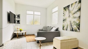Ein modernes Wohnzimmer mit grauem Sofa, weißem Hocker, rundem Couchtisch und einem an der Wand montierten Flachbildfernseher. Zwei Fenster mit Fensterläden, ein Deckenventilator und ein großes abstraktes Gemälde zieren die Wand.