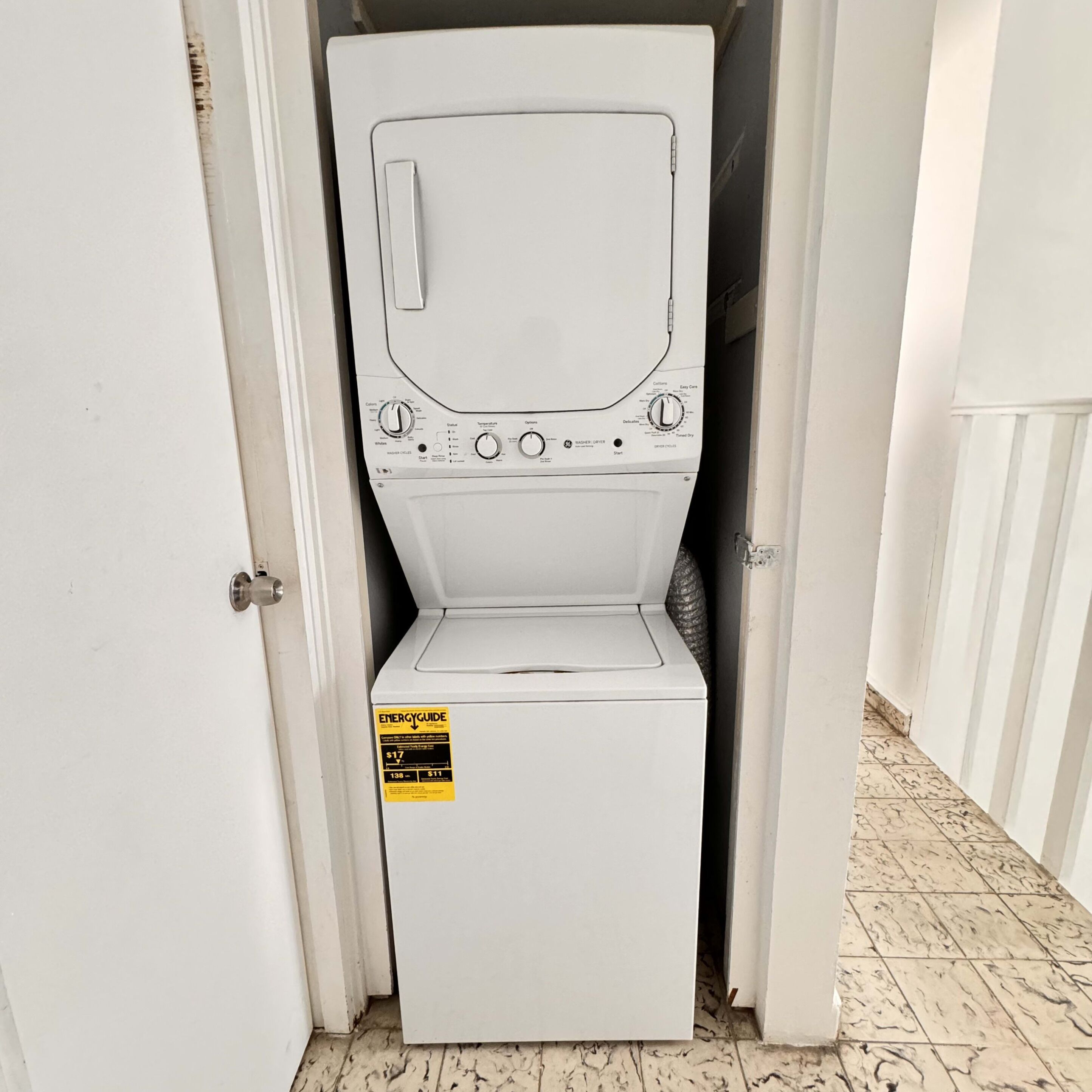 A compact, white washing machine in a narrow hallway with a door on one side.