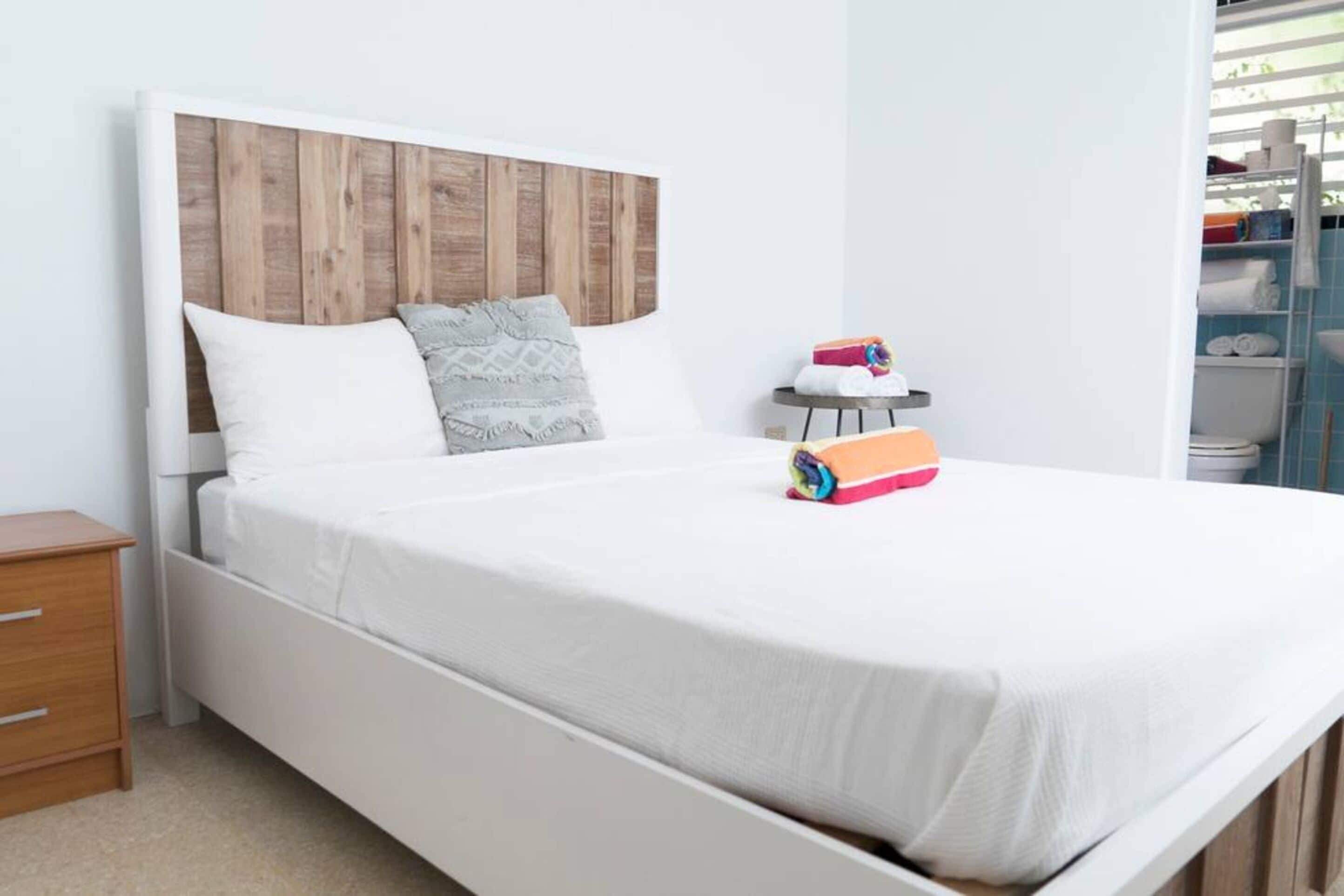 Ein ordentlich bezogenes Bett mit Holz Kopfteil, ein Nachttisch mit Lampe und ein kleines Regal mit Handtüchern.