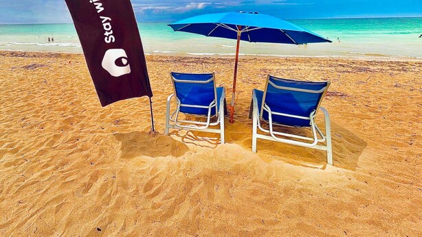 Ein Strand mit zwei blauen Stühlen, einem blauen Sonnenschirm und einer schwarzen Flagge mit Text.