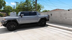 Un camionnette argentée est garée sur une surface en béton, avec un mur en béton et des arbres en arrière-plan.