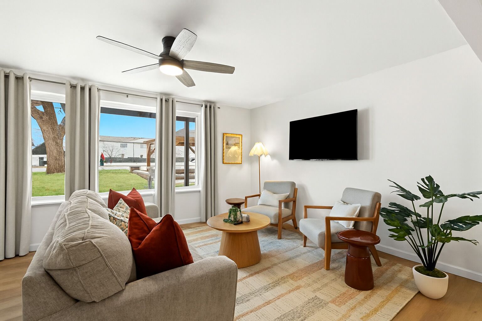 A modern living room with a sofa, chairs, a coffee table, a ceiling fan, a TV, and a large window with curtains.