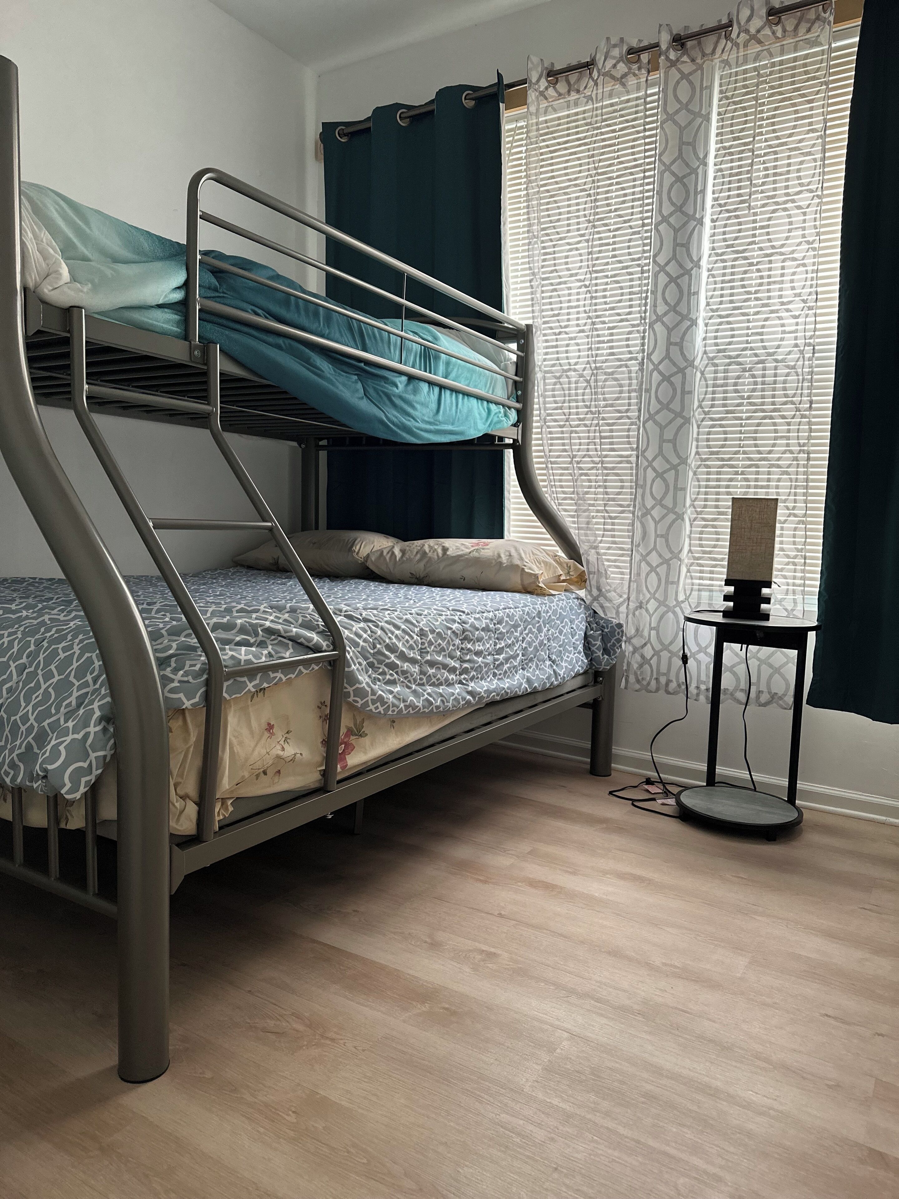 Una cama litera con escritorio y silla, una ventana con cortinas estampadas, y una mesita de noche con lámpara.