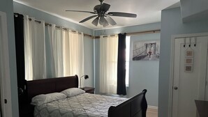 A bedroom with a bed, a ceiling fan, a nightstand, a door with a coat rack, and a wall with a framed picture.