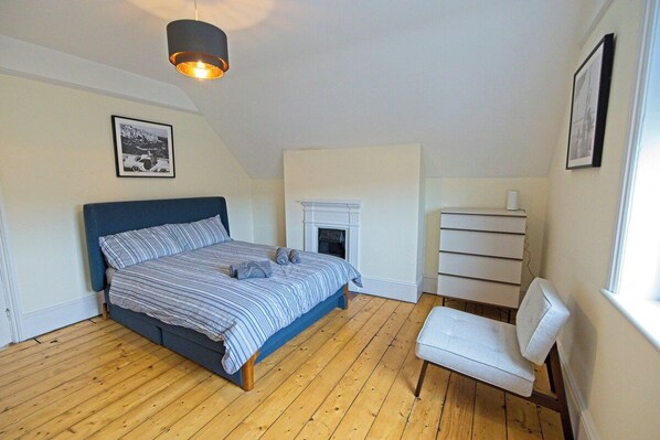 A bedroom with a bed, a white chair, a fireplace, and a picture on the wall.