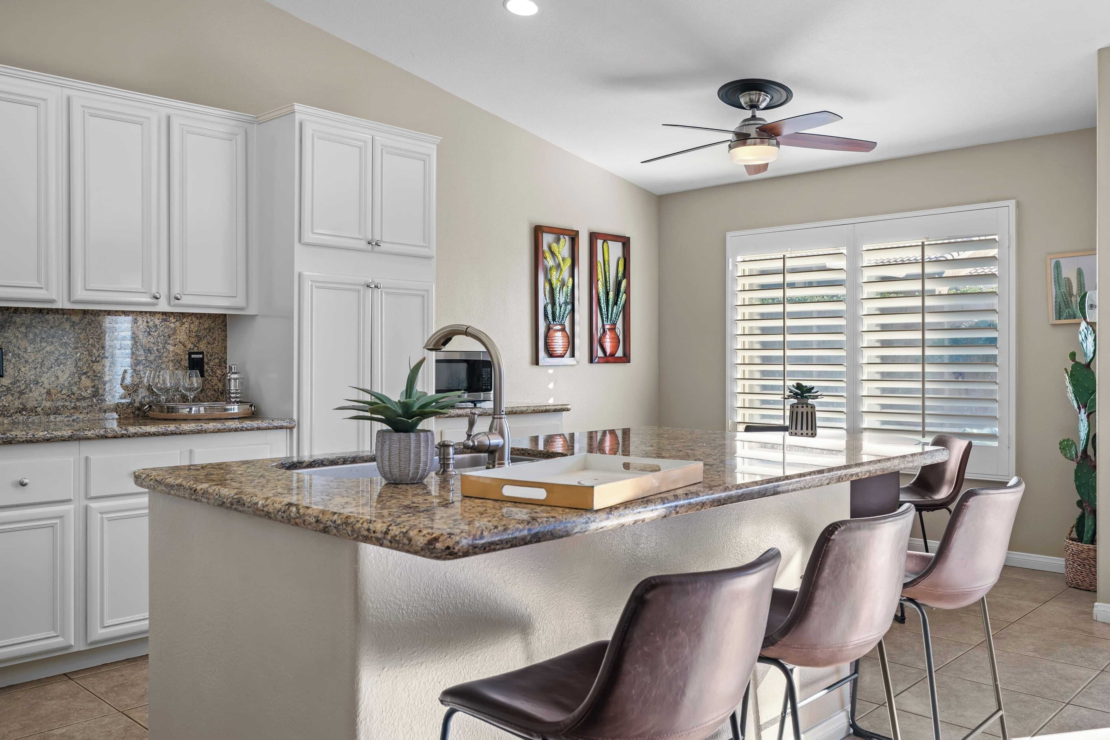 A kitchen with white cabinets, granite countertops, a central island with bar stools, a ceiling fan, and a window with blinds.