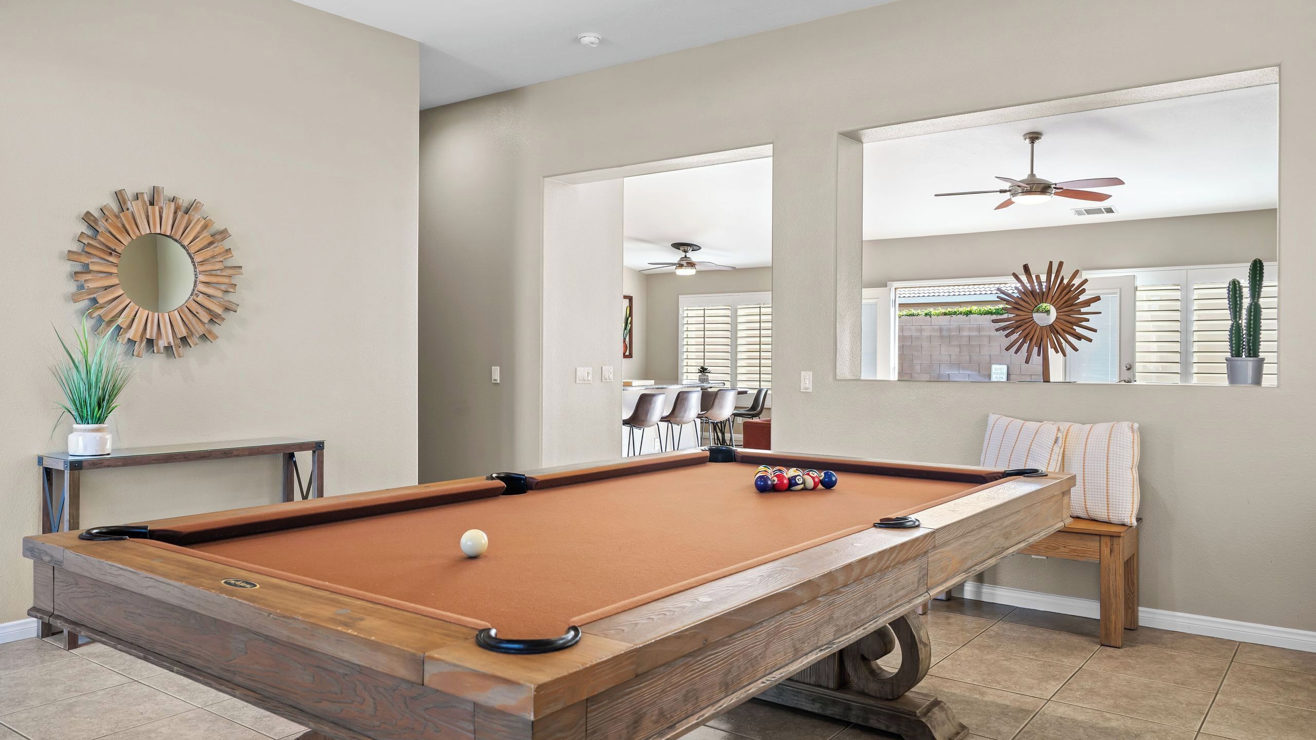 A pool table with balls, a sunburst mirror, and a ceiling fan in a room with a view of a dining area.