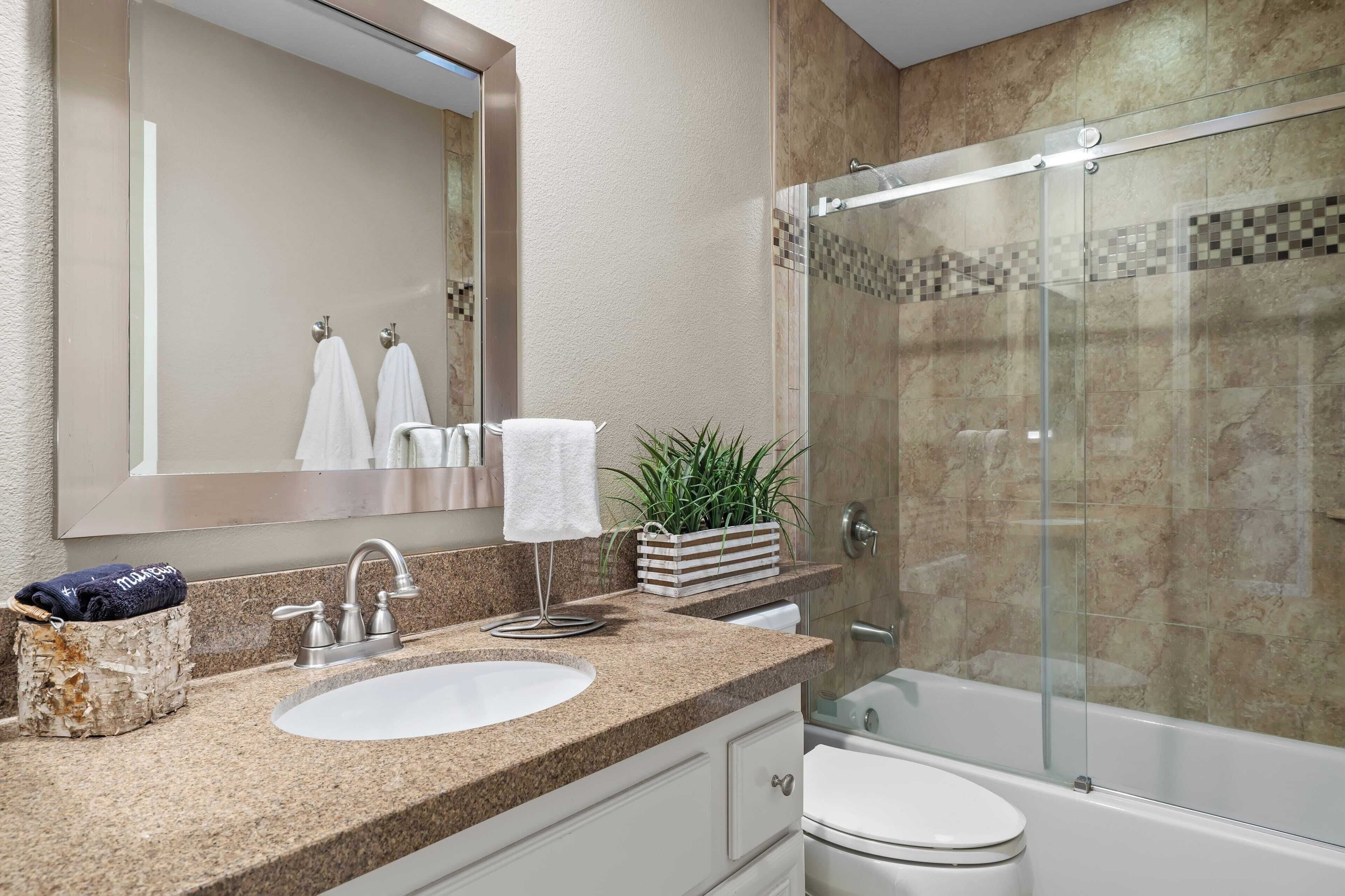 A modern bathroom with a granite countertop, a white sink, a large mirror, a glass-enclosed shower, and a toilet.