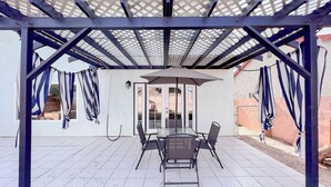 Ein überdachter Patio-Bereich mit Tisch und Stühlen, einer Pergola und Blick auf ein Haus durch die offene Tür.