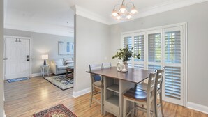 A dining area with a wooden table and chairs, a chandelier, and a view through white shutters. - Beautifully Decorated Condo, St. Simons Island, 8 minutes to Beach, Heated Pool (St. Simons Island)