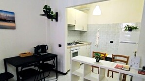 A compact kitchen with white cabinets, a stove, and a sink. There is a dining table with chairs, a small plant, and a clock on the wall.