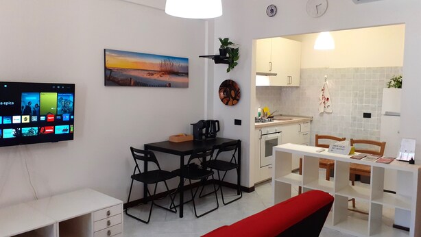 A modern living room with a flat-screen TV, a dining area with chairs, a red sofa, and a kitchen with white cabinets.