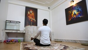 A person sitting on a patterned rug in a room with a large painting of a meditating figure on the wall, a marble bench, and a row of colorful yoga mats.