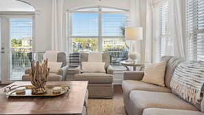 Ein Wohnzimmer mit einer Couch, Sesseln, einem Couchtisch und einer Lampe. Große Fenster mit Jalousien, Blick ins Freie und eine dekorative Holzkonstruktion auf dem Couchtisch.