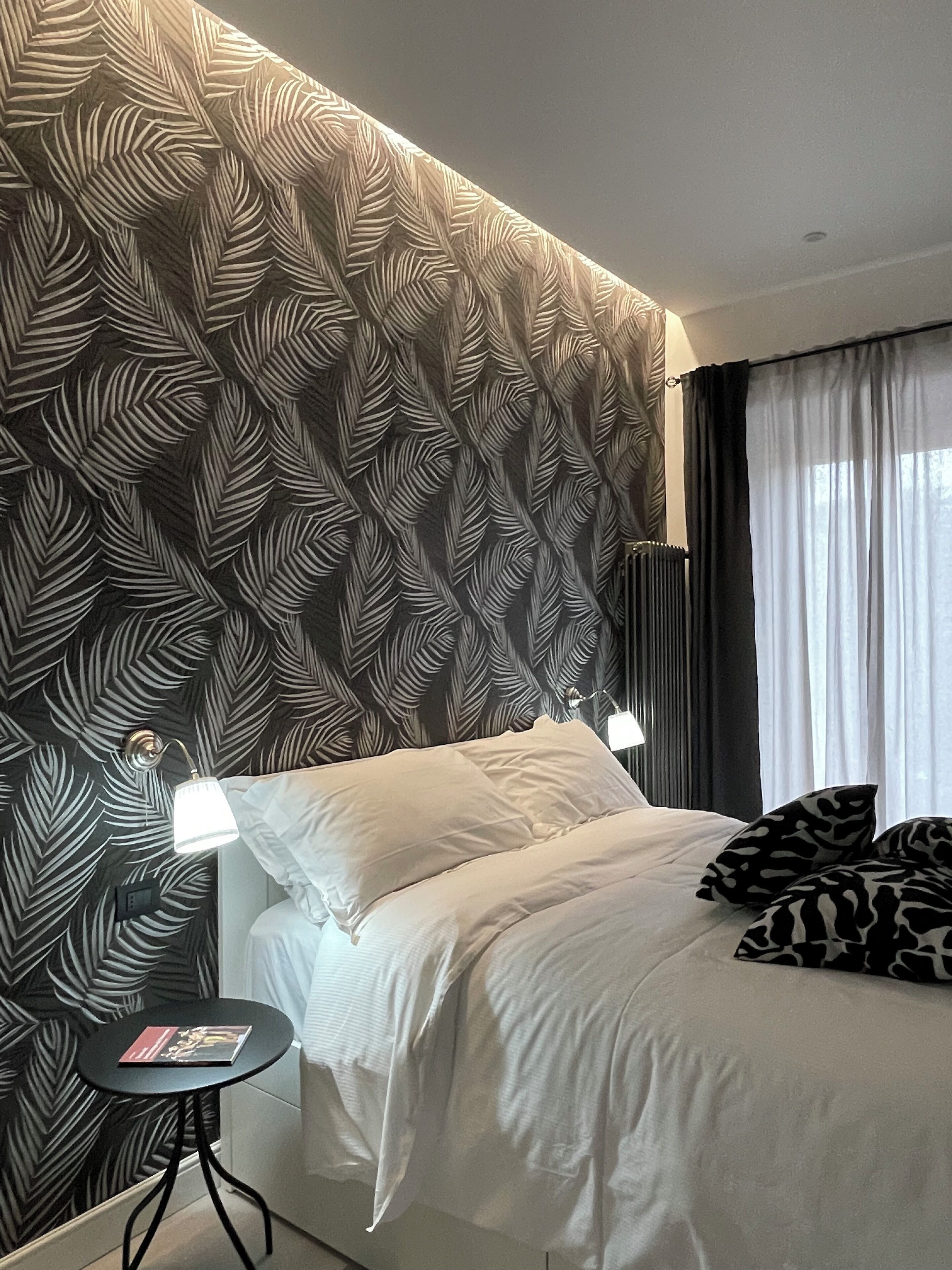 Una camera d'albergo con una testiera a motivi decorativi, un letto con lenzuola bianche, due abat-jour, un tavolino rotondo con un libro e una finestra con tende.