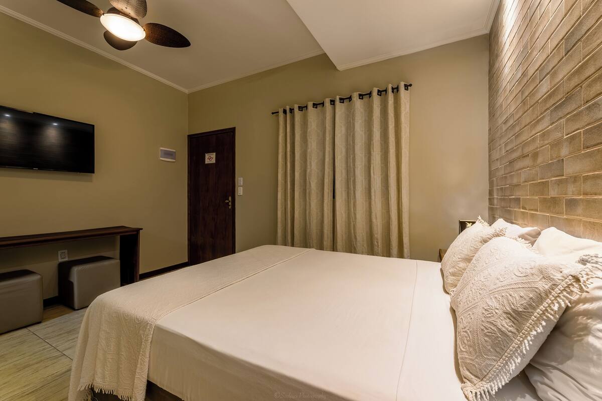 Une chambre d’hôtel équipée d’un lit, d’un téléviseur à écran plat fixé au mur, d’un ventilateur de plafond et d’une fenêtre avec des rideaux.