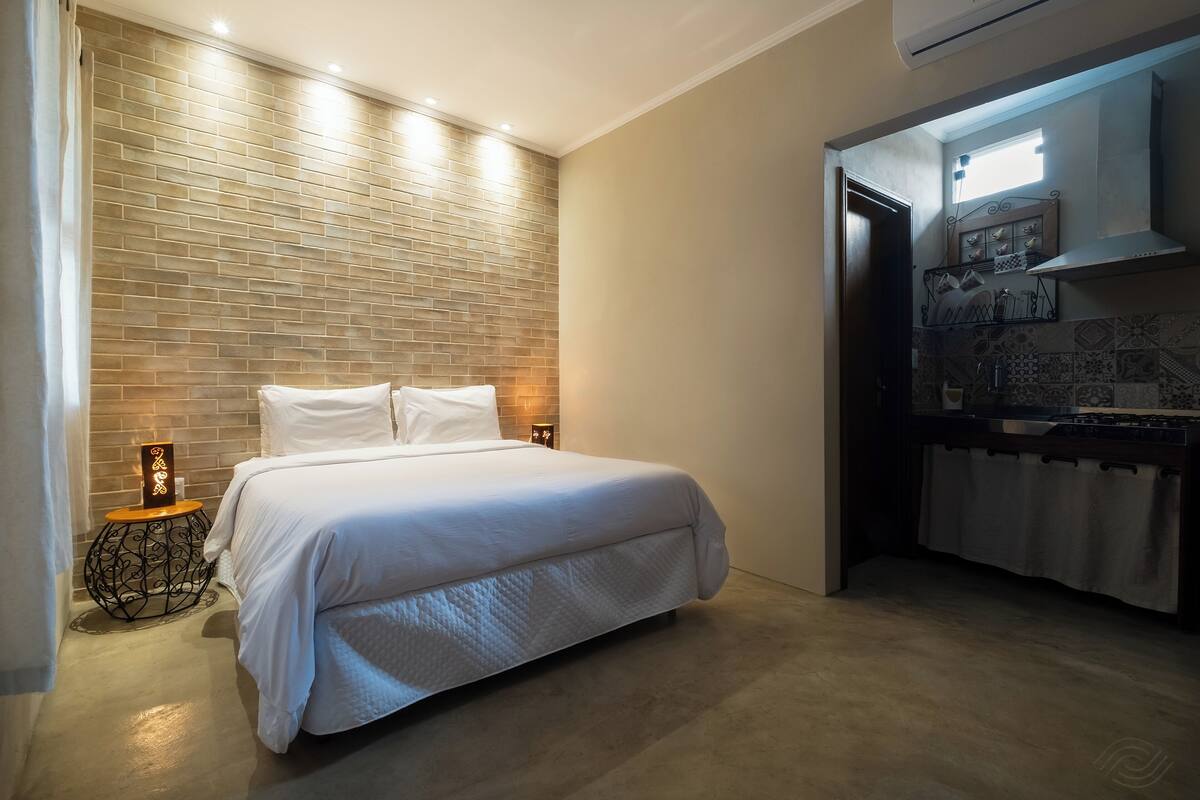 Une chambre avec un grand lit, un mur décoré d’un motif en briques, une petite table de chevet avec une lampe, et un coin cuisine avec une fenêtre au-dessus de la cuisinière.