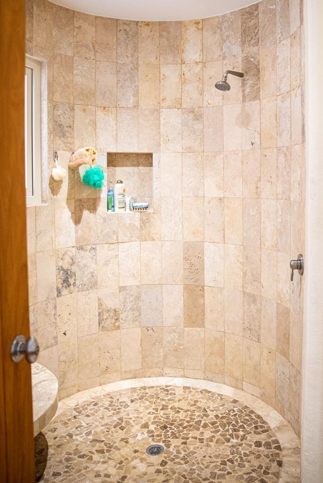 A shower with beige tiles, a drain, and a shelf with toiletries.