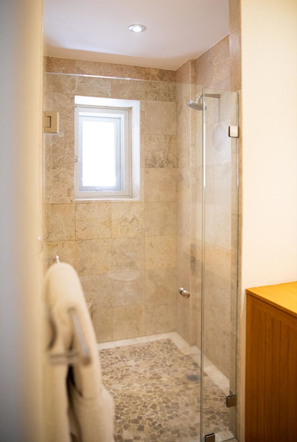 Una ducha con cerramiento de vidrio, una ventana pequeña, suelo de baldosas y un mueble de madera.