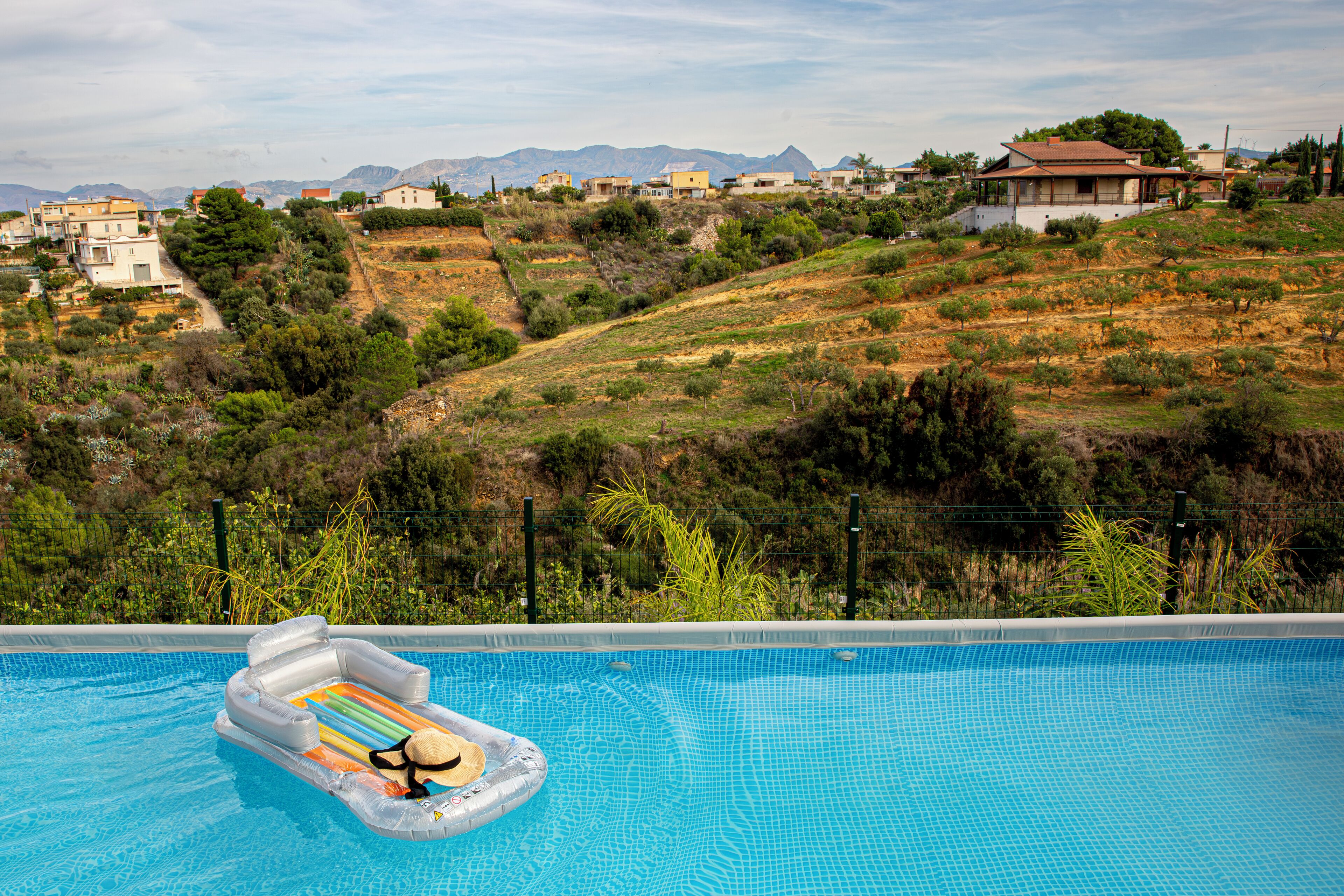 A swimming pool with a floating raft, surrounded by a hilly landscape with houses and vegetation.