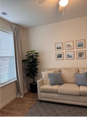 A living room with a beige sofa, a potted plant, and framed pictures on the wall.