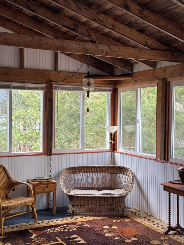 A cozy room with a wicker sofa, a small table, and a lamp. Large windows offer a view of trees outside.