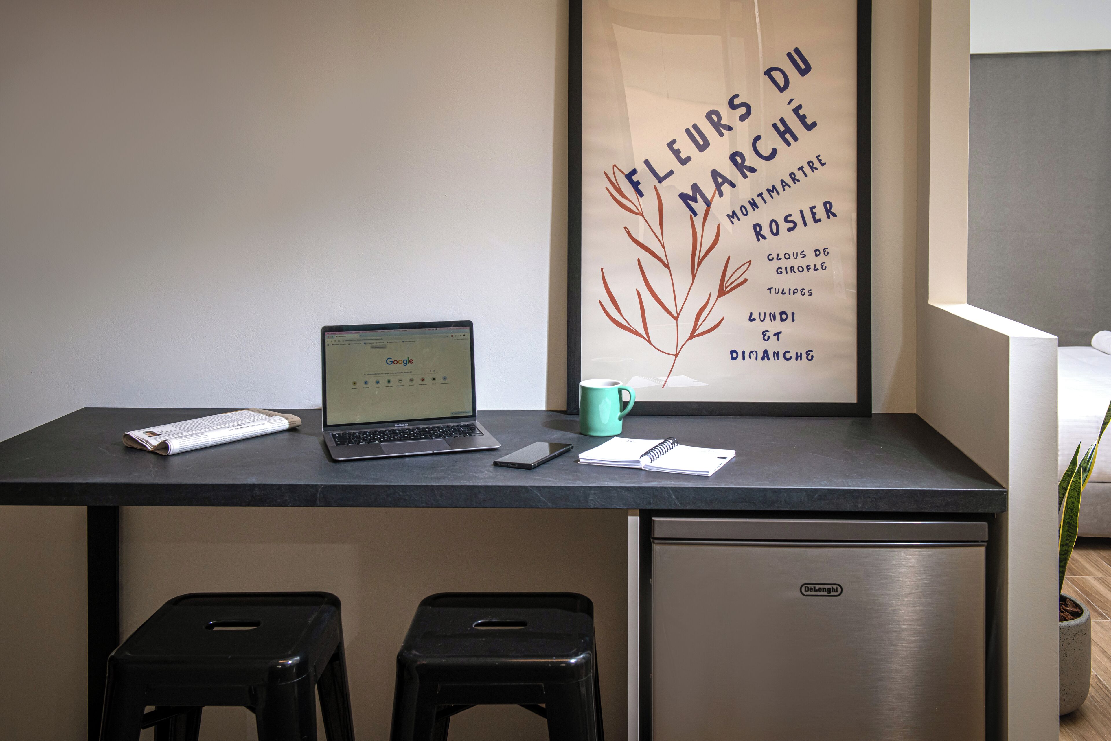 Un'area lavoro con un laptop, uno smartphone, un quaderno e una tazza su una scrivania. Sotto la scrivania, si trova un frigorifero e due sgabelli neri. Sullo sfondo, si intravede un poster incorniciato con del testo e una pianta.