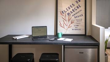 Un'area lavoro con un laptop, uno smartphone, un quaderno e una tazza su una scrivania. Sotto la scrivania, si trova un frigorifero e due sgabelli neri. Sullo sfondo, si intravede un poster incorniciato con del testo e una pianta.