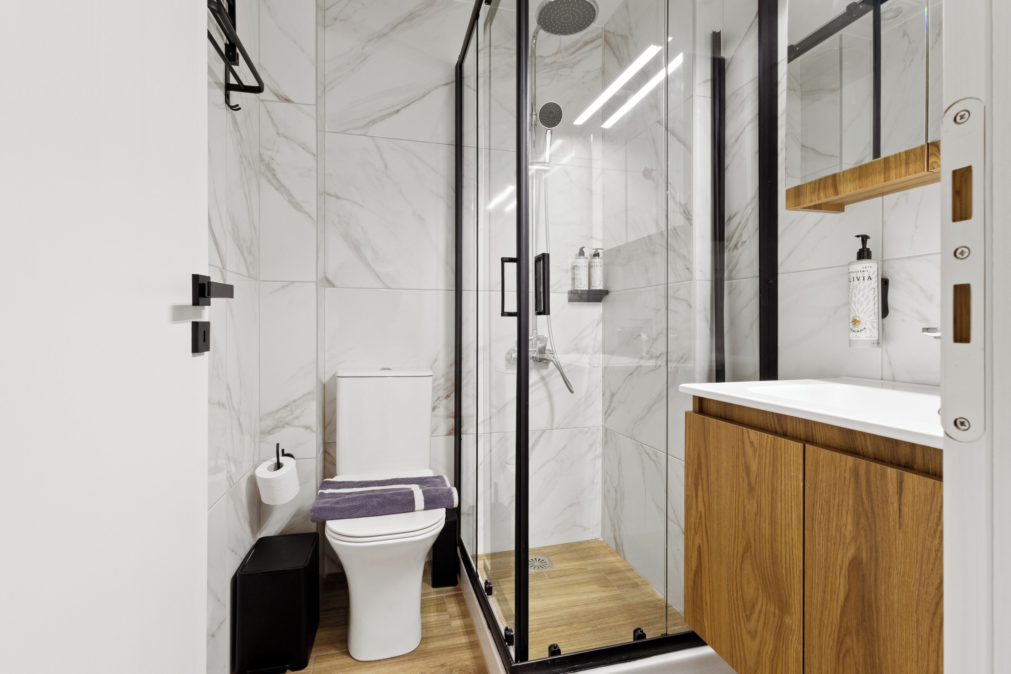 A modern bathroom with a glass-enclosed shower, a toilet, a sink with a wooden cabinet, and a mirror with a wooden shelf above it.