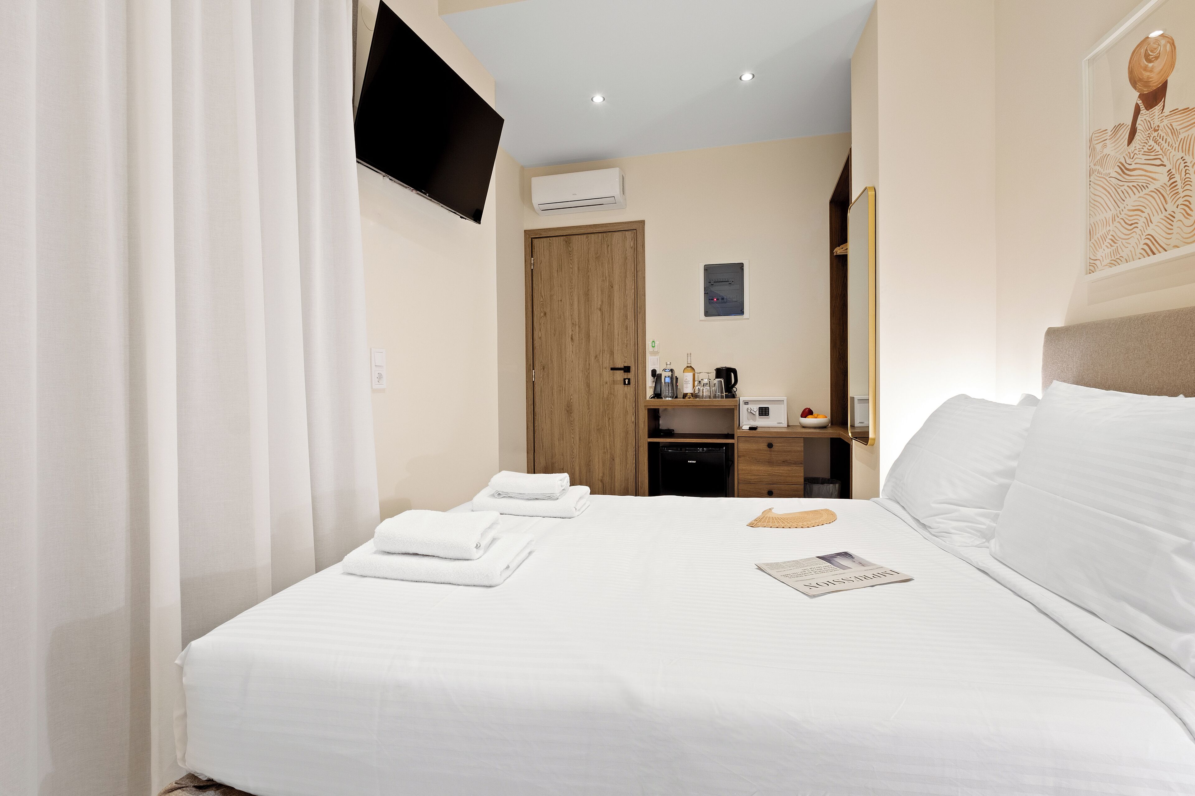 A hotel room with a neatly made bed, a flat-screen TV mounted on the wall, a wooden door, a small desk with a coffee maker, and a framed picture on the wall.
