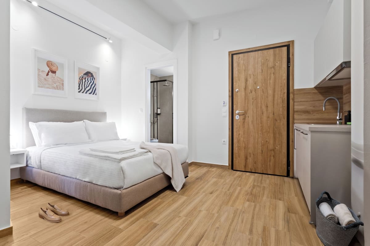 Une chambre moderne avec un lit, une porte en bois, une petite kitchenette et une salle de bain visible à travers une porte ouverte.