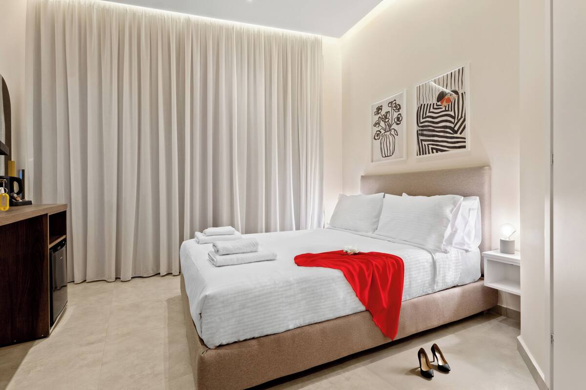 Une chambre d’hôtel moderne avec un grand lit, du linge de lit blanc, un plaid rouge et une paire de chaussures posées sur le sol.