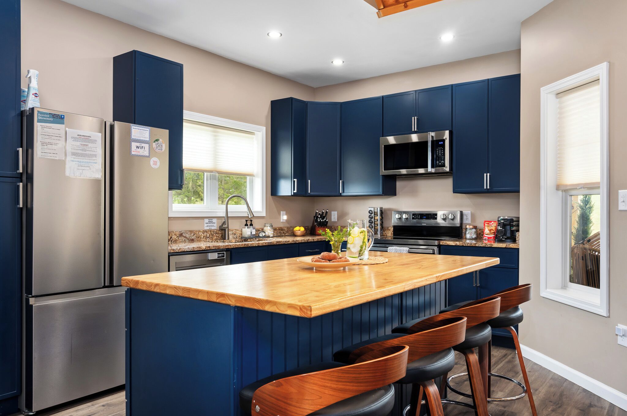 A modern kitchen with dark blue cabinets, stainless steel appliances, a wooden island with bar stools, and a window providing natural light.