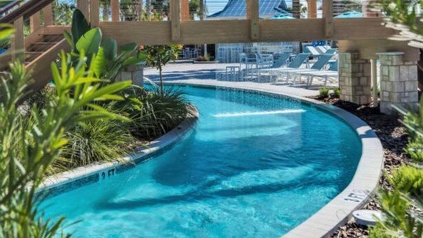 Une piscine dotée d’une terrasse en bois et de chaises longues, entourée de palmiers et offrant une vue sur un bâtiment dont le toit est en pente.