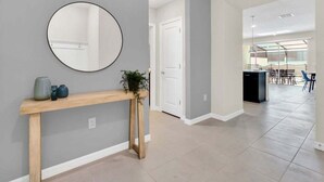 Un intérieur moderne avec un mur gris, un miroir rond, une console en bois agrémentée d’objets décoratifs, et une vue sur un coin repas avec des chaises et une table.