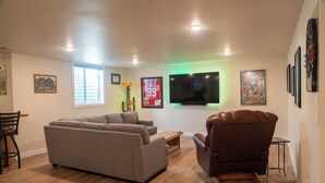 A modern living room with a grey sectional sofa, a brown leather armchair, and a flat-screen TV mounted on the wall.
